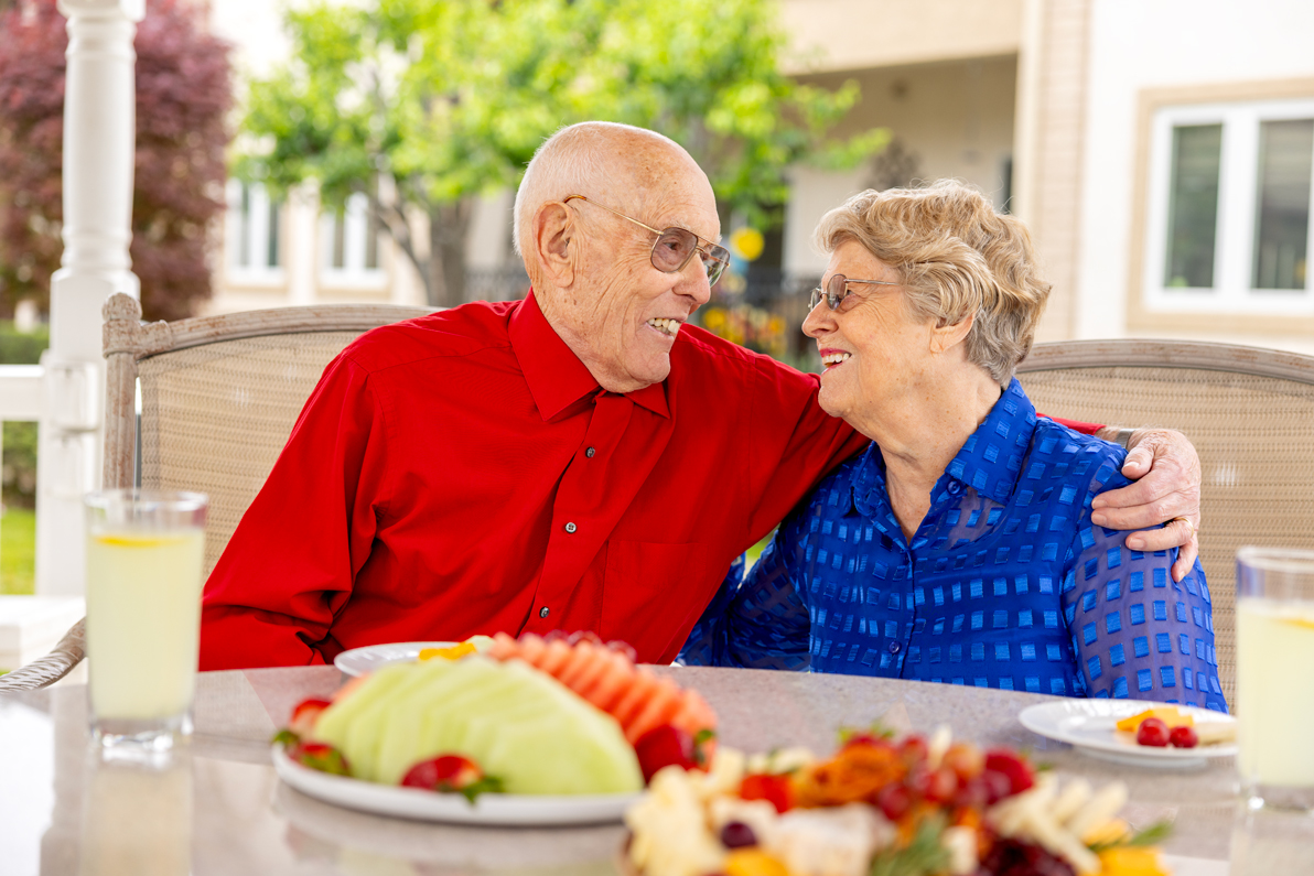 elderly male resident hugging elderly female resident at a table outdoors